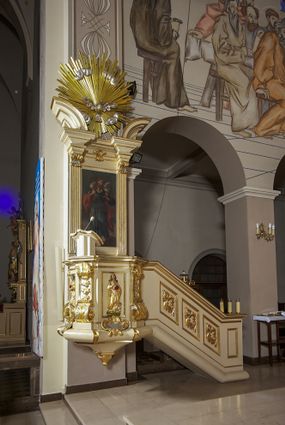 Zdjęcie nr 1: Ambona przyścienna, wisząca, dostępna schodami z podestem. Ich balustrada jest pełna dekorowana płycinami wypełnionymi plakietami z ornamentacją roślinną. Kosz na profilowanym cokole z półwałkiem, na planie prostokąta o wklęsło-wypukłych ściankach wydzielonych spływami z wolutami, dekorowanymi kampanulami i liśćmi akantu. Na wklęsłych ściankach płyciny w kształcie stojącego prostokąta, wypełnione płaskorzeźbionymi, całopostaciowymi przedstawieniami św. Łukasza z lewej i św. Jana Ewangelisty z prawej strony, pod ich stopami kartusze ujęte dekoracją rocaille’ową.  Parapet w postaci profilowanego gzymsu wyłamanego nad spływami. Spód kosza w kształcie wklęsło-wypukłego, odwróconego stożka zakończonego szyszką. Zaplecek w formie wydłużonego prostokąta ujętego kanelowanymi pilastrami. W polu obraz w kształcie stojącego prostokąta z wizerunkiem Chrystusa Dobrego Pasterza. Pilastry podtrzymują profilowane belkowanie wyłamane na ich osiach, na których znajdują się fragmenty przyczółka skierowane na zewnątrz. Między nimi, na ścianie rozbudowane przedstawienie gołębicy Ducha Świętego na tle glorii promienistej  i obłoków.  Struktura w odcieniu kremowym, dekoracja i profile złocone i srebrzone. 