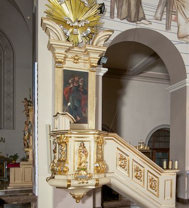 Zdjęcie nr 2: Ambona przyścienna, wisząca, dostępna schodami z podestem. Ich balustrada jest pełna dekorowana płycinami wypełnionymi plakietami z ornamentacją roślinną. Kosz na profilowanym cokole z półwałkiem, na planie prostokąta o wklęsło-wypukłych ściankach wydzielonych spływami z wolutami, dekorowanymi kampanulami i liśćmi akantu. Na wklęsłych ściankach płyciny w kształcie stojącego prostokąta, wypełnione płaskorzeźbionymi, całopostaciowymi przedstawieniami św. Łukasza z lewej i św. Jana Ewangelisty z prawej strony, pod ich stopami kartusze ujęte dekoracją rocaille’ową.  Parapet w postaci profilowanego gzymsu wyłamanego nad spływami. Spód kosza w kształcie wklęsło-wypukłego, odwróconego stożka zakończonego szyszką. Zaplecek w formie wydłużonego prostokąta ujętego kanelowanymi pilastrami. W polu obraz w kształcie stojącego prostokąta z wizerunkiem Chrystusa Dobrego Pasterza. Pilastry podtrzymują profilowane belkowanie wyłamane na ich osiach, na których znajdują się fragmenty przyczółka skierowane na zewnątrz. Między nimi, na ścianie rozbudowane przedstawienie gołębicy Ducha Świętego na tle glorii promienistej  i obłoków.  Struktura w odcieniu kremowym, dekoracja i profile złocone i srebrzone. 