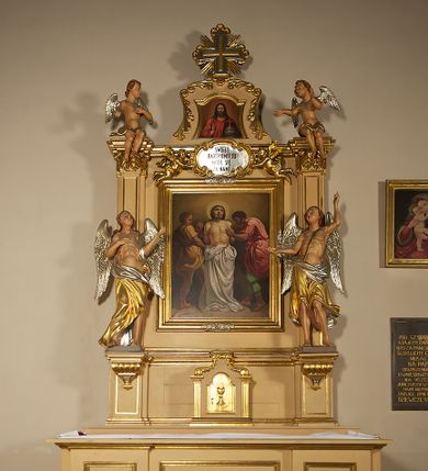 Zdjęcie nr 1: Ołtarz przyścienny, architektoniczny, jednoosiowy, jednokondygnacyjny ze zwieńczeniem. Mensa prostopadłościenna na profilowanym cokole, antepedium dekorowane trzema płycinami z plakietami z ornamentu roślinnego. Tabernakulum architektoniczne, w formie prostopadłościanu na profilowanym cokole, o ujętym wolutami wklęsło-wypukłym zwieńczeniu. Na ścianie frontowej drzwiczki w kształcie stojącego prostokąta zamkniętego łukiem nadwieszonym z uskokiem. Nastawa ołtarzowa ujęta  pilastrami z płycinami ustawionymi na wysokich cokołach, na których wspierają się konsole podtrzymujące rzeźby aniołów. Podpory dźwigają pełne, wyłamane na ich osiach belkowanie, na którym posadowiono figury aniołków. W centrum kondygnacji prostokątna nisza mieszcząca obraz „Męczeństwo św. Bartłomieja”, ujęta prostokątną ramą dekorowaną w czterech miejscach kwitnącymi różami z liśćmi. Między polem obrazowym a belkowaniem kondygnacji unoszony przez dwa anioły kartusz z inskrypcją „ŚWIĘTY / BARTŁOMIEJU / MÓDL SIĘ / ZA NAMI”. Zwieńczenie o kształcie wklęsło-wypukłym, ujęte ulistnionymi wolutami, wypełnione obrazem powtarzającym jego kształt, przedstawiającym Jezusa Chrystusa. Na szczycie krzyż równoramienny o ramionach zakończonych trójlistnie, na tle glorii.  Struktura ołtarza w kolorze kremowym; profile, detale i ornamenty złocone i srebrzone.