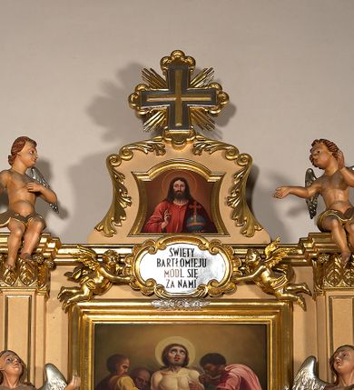 Zdjęcie nr 4: Ołtarz przyścienny, architektoniczny, jednoosiowy, jednokondygnacyjny ze zwieńczeniem. Mensa prostopadłościenna na profilowanym cokole, antepedium dekorowane trzema płycinami z plakietami z ornamentu roślinnego. Tabernakulum architektoniczne, w formie prostopadłościanu na profilowanym cokole, o ujętym wolutami wklęsło-wypukłym zwieńczeniu. Na ścianie frontowej drzwiczki w kształcie stojącego prostokąta zamkniętego łukiem nadwieszonym z uskokiem. Nastawa ołtarzowa ujęta  pilastrami z płycinami ustawionymi na wysokich cokołach, na których wspierają się konsole podtrzymujące rzeźby aniołów. Podpory dźwigają pełne, wyłamane na ich osiach belkowanie, na którym posadowiono figury aniołków. W centrum kondygnacji prostokątna nisza mieszcząca obraz „Męczeństwo św. Bartłomieja”, ujęta prostokątną ramą dekorowaną w czterech miejscach kwitnącymi różami z liśćmi. Między polem obrazowym a belkowaniem kondygnacji unoszony przez dwa anioły kartusz z inskrypcją „ŚWIĘTY / BARTŁOMIEJU / MÓDL SIĘ / ZA NAMI”. Zwieńczenie o kształcie wklęsło-wypukłym, ujęte ulistnionymi wolutami, wypełnione obrazem powtarzającym jego kształt, przedstawiającym Jezusa Chrystusa. Na szczycie krzyż równoramienny o ramionach zakończonych trójlistnie, na tle glorii.  Struktura ołtarza w kolorze kremowym; profile, detale i ornamenty złocone i srebrzone.