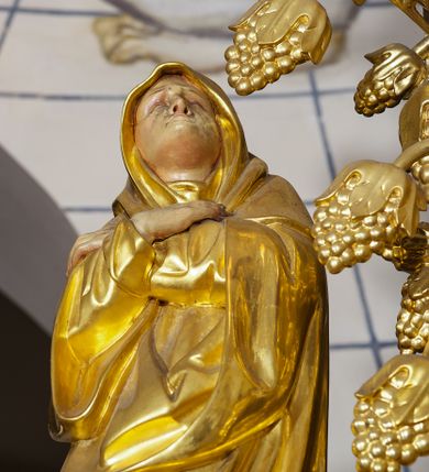 Zdjęcie nr 26: Ołtarz w formie rzeźbionej, ażurowej ramy na trójstopniowym cokole, z prostopadłościenną mensą z antepedium dekorowanym kwadratowymi płycinami z motywem czworoliścia. Na nim prostopadłościenne tabernakulum z drzwiami w kształcie stojącego prostokąta dekorowanymi koszem z chlebem, rybami i chrystogramem. Środkowa część podstawy wypełniona płycinami w kształcie leżących prostokątów z plakietami roślinnymi. Na jej zewnętrznych częściach posadowiono figury klęczących aniołów. Najwyższy stopień węższy, trójosiowy, o osiach zewnętrznych wysuniętych, flankowany konsolami z rzeźbami Matki Boskiej po lewej stronie i św. Jana Ewangelisty po prawej. Centralnie umieszczona rama ujmuje pole w kształcie serca, na tle którego jest eksponowany krucyfiks. Ażur tworzy wić winnej latorośli z kiśćmi winogron, a w nią wkomponowane jest siedem postaci aniołów trzymających banderole z ostatnimi słowami wypowiedzianymi przez Chrystusa wiszącego na krzyżu. Od góry: „Pragnę”, „Niewiasto! Oto syn Twój”, „Oto matka twoja”, „Ojcze odpuść im, / bo nie wiedzą co czynią”, „Zaprawdę powiadam tobie / dziś jeszcze będziesz ze Mną w raju”, „Spełniło się! / Ojcze w ręce Twoje oddaję ducha mojego”, „Boże mój Boże mój / czemużeś mnie opuścił”. Struktura w kolorze kremowym, profile, detale i ornamenty złocone; banderole srebrzone. 