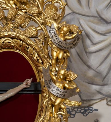 Zdjęcie nr 10: Ołtarz w formie rzeźbionej, ażurowej ramy na trójstopniowym cokole, z prostopadłościenną mensą z antepedium dekorowanym kwadratowymi płycinami z motywem czworoliścia. Na nim prostopadłościenne tabernakulum z drzwiami w kształcie stojącego prostokąta dekorowanymi koszem z chlebem, rybami i chrystogramem. Środkowa część podstawy wypełniona płycinami w kształcie leżących prostokątów z plakietami roślinnymi. Na jej zewnętrznych częściach posadowiono figury klęczących aniołów. Najwyższy stopień węższy, trójosiowy, o osiach zewnętrznych wysuniętych, flankowany konsolami z rzeźbami Matki Boskiej po lewej stronie i św. Jana Ewangelisty po prawej. Centralnie umieszczona rama ujmuje pole w kształcie serca, na tle którego jest eksponowany krucyfiks. Ażur tworzy wić winnej latorośli z kiśćmi winogron, a w nią wkomponowane jest siedem postaci aniołów trzymających banderole z ostatnimi słowami wypowiedzianymi przez Chrystusa wiszącego na krzyżu. Od góry: „Pragnę”, „Niewiasto! Oto syn Twój”, „Oto matka twoja”, „Ojcze odpuść im, / bo nie wiedzą co czynią”, „Zaprawdę powiadam tobie / dziś jeszcze będziesz ze Mną w raju”, „Spełniło się! / Ojcze w ręce Twoje oddaję ducha mojego”, „Boże mój Boże mój / czemużeś mnie opuścił”. Struktura w kolorze kremowym, profile, detale i ornamenty złocone; banderole srebrzone. 