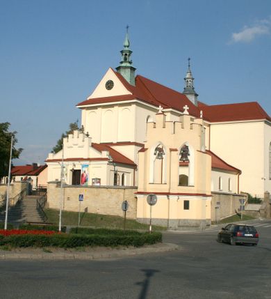 Zdjęcie nr 144: Kościół murowany, orientowany, jednonawowy, z transeptem, prezbiterium z obejściem i przybudówkami. Od zachodu kruchta, od północy kwadratowa kaplica, od południa kolejna przybudówka. 
Prezbiterium zamknięte półokrągło, z obejściem przeprutym do niego arkadami wspartymi na filarach. Ściany nawy i prezbiterium gładkie, przekryte sklepieniem kolebkowym z lunetami, w których mieszczą się okna. Transept i przecięcie naw sklepione krzyżowo. Na ścianach polichromia figuralna ze scenami z Nowego Testamentu, w partii sklepienia drobne, złote gwiazdki. Od zachodu chór zatopiony w występującym asymetrycznie murze, z tralkową balustradą. W prezbiterium siedem okien w kształcie stojącego prostokąta zamkniętego łukiem pełnym wypełnionych witrażami, w nawie sześć okien, w ramionach transeptu po jednym, czterodzielnym, wypełnionym witrażem, na wysokości chóru po dwa z każdej strony. Główne wejście od zachodu przez kruchtę. Fasada poprzedzona przedsionkiem o podziałach ramowych, z prostokątnym wejściem na osi, flankowanym parą pilastrów i prostokątnymi płycinami. Kondygnację wieńczy pełne belkowanie z wysuniętym gzymsem podtrzymującym daszek i trójkątny, schodkowy szczyt zakończony postumentami. Na centralnym krzyż, na sąsiadujących figury dzieci z rogami obfitości, a na zewnętrznych wazony płomieniste; szczyt ujmują postumenty z kulami. Fasada dwukondygnacyjna ze szczytem. Kondygnacja trójosiowa, rozdzielona pilastrami, o polach wypełnionych blendami w kształcie stojących prostokątów zamkniętych łukiem pełnym; szczyt trójkątny, z tarczą zegarową pośrodku. Budynek kościoła o wyraźnych podziałach na bryły składowe, ściany gładkie, przeprute oknami, z gzymsami pod krawędziami dachu. 
Nad nawą główną, na kalenicy, wieżyczka na sygnaturkę z latarnią i baniastym hełmem, druga na skrzyżowaniu naw, z latarnią i  hełmem zakończonym krzyżem. Dach nad nawą główną i przedsionkiem dwuspadowy, nad ramionami transeptu trójspadowym,nad prezbiterium zakończony wielobocznie, na przybudówkach dachy pulpitowe, dwuspadowe i wielospadowe. Dachy kryte dachówką ceramiczną, wieżyczki blachą. 
Najciekawszymi zabytkami kościoła są ołtarz główny z około 1700 roku z nastawą utworzoną z wici winnej latorośli, manierystyczny ołtarz boczny oraz siedemnastowieczna chrzcielnica z warsztatu Santi Gucciego. 
