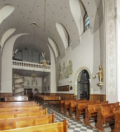 Zdjęcie nr 90: Kościół murowany, orientowany, jednonawowy, z transeptem, prezbiterium z obejściem i przybudówkami. Od zachodu kruchta, od północy kwadratowa kaplica, od południa kolejna przybudówka. 
Prezbiterium zamknięte półokrągło, z obejściem przeprutym do niego arkadami wspartymi na filarach. Ściany nawy i prezbiterium gładkie, przekryte sklepieniem kolebkowym z lunetami, w których mieszczą się okna. Transept i przecięcie naw sklepione krzyżowo. Na ścianach polichromia figuralna ze scenami z Nowego Testamentu, w partii sklepienia drobne, złote gwiazdki. Od zachodu chór zatopiony w występującym asymetrycznie murze, z tralkową balustradą. W prezbiterium siedem okien w kształcie stojącego prostokąta zamkniętego łukiem pełnym wypełnionych witrażami, w nawie sześć okien, w ramionach transeptu po jednym, czterodzielnym, wypełnionym witrażem, na wysokości chóru po dwa z każdej strony. Główne wejście od zachodu przez kruchtę. Fasada poprzedzona przedsionkiem o podziałach ramowych, z prostokątnym wejściem na osi, flankowanym parą pilastrów i prostokątnymi płycinami. Kondygnację wieńczy pełne belkowanie z wysuniętym gzymsem podtrzymującym daszek i trójkątny, schodkowy szczyt zakończony postumentami. Na centralnym krzyż, na sąsiadujących figury dzieci z rogami obfitości, a na zewnętrznych wazony płomieniste; szczyt ujmują postumenty z kulami. Fasada dwukondygnacyjna ze szczytem. Kondygnacja trójosiowa, rozdzielona pilastrami, o polach wypełnionych blendami w kształcie stojących prostokątów zamkniętych łukiem pełnym; szczyt trójkątny, z tarczą zegarową pośrodku. Budynek kościoła o wyraźnych podziałach na bryły składowe, ściany gładkie, przeprute oknami, z gzymsami pod krawędziami dachu. 
Nad nawą główną, na kalenicy, wieżyczka na sygnaturkę z latarnią i baniastym hełmem, druga na skrzyżowaniu naw, z latarnią i  hełmem zakończonym krzyżem. Dach nad nawą główną i przedsionkiem dwuspadowy, nad ramionami transeptu trójspadowym,nad prezbiterium zakończony wielobocznie, na przybudówkach dachy pulpitowe, dwuspadowe i wielospadowe. Dachy kryte dachówką ceramiczną, wieżyczki blachą. 
Najciekawszymi zabytkami kościoła są ołtarz główny z około 1700 roku z nastawą utworzoną z wici winnej latorośli, manierystyczny ołtarz boczny oraz siedemnastowieczna chrzcielnica z warsztatu Santi Gucciego. 
