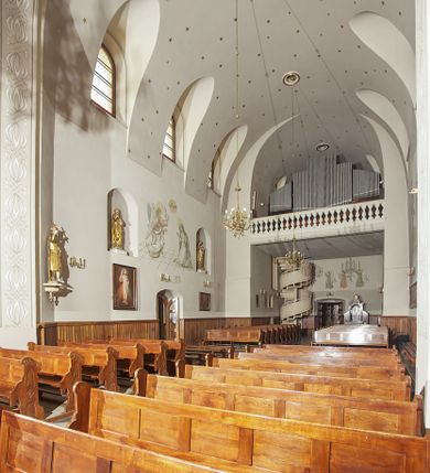 Zdjęcie nr 113: Kościół murowany, orientowany, jednonawowy, z transeptem, prezbiterium z obejściem i przybudówkami. Od zachodu kruchta, od północy kwadratowa kaplica, od południa kolejna przybudówka. 
Prezbiterium zamknięte półokrągło, z obejściem przeprutym do niego arkadami wspartymi na filarach. Ściany nawy i prezbiterium gładkie, przekryte sklepieniem kolebkowym z lunetami, w których mieszczą się okna. Transept i przecięcie naw sklepione krzyżowo. Na ścianach polichromia figuralna ze scenami z Nowego Testamentu, w partii sklepienia drobne, złote gwiazdki. Od zachodu chór zatopiony w występującym asymetrycznie murze, z tralkową balustradą. W prezbiterium siedem okien w kształcie stojącego prostokąta zamkniętego łukiem pełnym wypełnionych witrażami, w nawie sześć okien, w ramionach transeptu po jednym, czterodzielnym, wypełnionym witrażem, na wysokości chóru po dwa z każdej strony. Główne wejście od zachodu przez kruchtę. Fasada poprzedzona przedsionkiem o podziałach ramowych, z prostokątnym wejściem na osi, flankowanym parą pilastrów i prostokątnymi płycinami. Kondygnację wieńczy pełne belkowanie z wysuniętym gzymsem podtrzymującym daszek i trójkątny, schodkowy szczyt zakończony postumentami. Na centralnym krzyż, na sąsiadujących figury dzieci z rogami obfitości, a na zewnętrznych wazony płomieniste; szczyt ujmują postumenty z kulami. Fasada dwukondygnacyjna ze szczytem. Kondygnacja trójosiowa, rozdzielona pilastrami, o polach wypełnionych blendami w kształcie stojących prostokątów zamkniętych łukiem pełnym; szczyt trójkątny, z tarczą zegarową pośrodku. Budynek kościoła o wyraźnych podziałach na bryły składowe, ściany gładkie, przeprute oknami, z gzymsami pod krawędziami dachu. 
Nad nawą główną, na kalenicy, wieżyczka na sygnaturkę z latarnią i baniastym hełmem, druga na skrzyżowaniu naw, z latarnią i  hełmem zakończonym krzyżem. Dach nad nawą główną i przedsionkiem dwuspadowy, nad ramionami transeptu trójspadowym,nad prezbiterium zakończony wielobocznie, na przybudówkach dachy pulpitowe, dwuspadowe i wielospadowe. Dachy kryte dachówką ceramiczną, wieżyczki blachą. 
Najciekawszymi zabytkami kościoła są ołtarz główny z około 1700 roku z nastawą utworzoną z wici winnej latorośli, manierystyczny ołtarz boczny oraz siedemnastowieczna chrzcielnica z warsztatu Santi Gucciego. 
