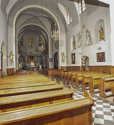 Zdjęcie nr 79: Kościół murowany, orientowany, jednonawowy, z transeptem, prezbiterium z obejściem i przybudówkami. Od zachodu kruchta, od północy kwadratowa kaplica, od południa kolejna przybudówka. 
Prezbiterium zamknięte półokrągło, z obejściem przeprutym do niego arkadami wspartymi na filarach. Ściany nawy i prezbiterium gładkie, przekryte sklepieniem kolebkowym z lunetami, w których mieszczą się okna. Transept i przecięcie naw sklepione krzyżowo. Na ścianach polichromia figuralna ze scenami z Nowego Testamentu, w partii sklepienia drobne, złote gwiazdki. Od zachodu chór zatopiony w występującym asymetrycznie murze, z tralkową balustradą. W prezbiterium siedem okien w kształcie stojącego prostokąta zamkniętego łukiem pełnym wypełnionych witrażami, w nawie sześć okien, w ramionach transeptu po jednym, czterodzielnym, wypełnionym witrażem, na wysokości chóru po dwa z każdej strony. Główne wejście od zachodu przez kruchtę. Fasada poprzedzona przedsionkiem o podziałach ramowych, z prostokątnym wejściem na osi, flankowanym parą pilastrów i prostokątnymi płycinami. Kondygnację wieńczy pełne belkowanie z wysuniętym gzymsem podtrzymującym daszek i trójkątny, schodkowy szczyt zakończony postumentami. Na centralnym krzyż, na sąsiadujących figury dzieci z rogami obfitości, a na zewnętrznych wazony płomieniste; szczyt ujmują postumenty z kulami. Fasada dwukondygnacyjna ze szczytem. Kondygnacja trójosiowa, rozdzielona pilastrami, o polach wypełnionych blendami w kształcie stojących prostokątów zamkniętych łukiem pełnym; szczyt trójkątny, z tarczą zegarową pośrodku. Budynek kościoła o wyraźnych podziałach na bryły składowe, ściany gładkie, przeprute oknami, z gzymsami pod krawędziami dachu. 
Nad nawą główną, na kalenicy, wieżyczka na sygnaturkę z latarnią i baniastym hełmem, druga na skrzyżowaniu naw, z latarnią i  hełmem zakończonym krzyżem. Dach nad nawą główną i przedsionkiem dwuspadowy, nad ramionami transeptu trójspadowym,nad prezbiterium zakończony wielobocznie, na przybudówkach dachy pulpitowe, dwuspadowe i wielospadowe. Dachy kryte dachówką ceramiczną, wieżyczki blachą. 
Najciekawszymi zabytkami kościoła są ołtarz główny z około 1700 roku z nastawą utworzoną z wici winnej latorośli, manierystyczny ołtarz boczny oraz siedemnastowieczna chrzcielnica z warsztatu Santi Gucciego. 
