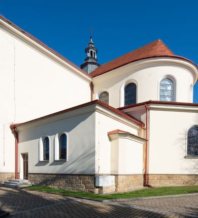 Zdjęcie nr 10: Kościół murowany, orientowany, jednonawowy, z transeptem, prezbiterium z obejściem i przybudówkami. Od zachodu kruchta, od północy kwadratowa kaplica, od południa kolejna przybudówka. 
Prezbiterium zamknięte półokrągło, z obejściem przeprutym do niego arkadami wspartymi na filarach. Ściany nawy i prezbiterium gładkie, przekryte sklepieniem kolebkowym z lunetami, w których mieszczą się okna. Transept i przecięcie naw sklepione krzyżowo. Na ścianach polichromia figuralna ze scenami z Nowego Testamentu, w partii sklepienia drobne, złote gwiazdki. Od zachodu chór zatopiony w występującym asymetrycznie murze, z tralkową balustradą. W prezbiterium siedem okien w kształcie stojącego prostokąta zamkniętego łukiem pełnym wypełnionych witrażami, w nawie sześć okien, w ramionach transeptu po jednym, czterodzielnym, wypełnionym witrażem, na wysokości chóru po dwa z każdej strony. Główne wejście od zachodu przez kruchtę. Fasada poprzedzona przedsionkiem o podziałach ramowych, z prostokątnym wejściem na osi, flankowanym parą pilastrów i prostokątnymi płycinami. Kondygnację wieńczy pełne belkowanie z wysuniętym gzymsem podtrzymującym daszek i trójkątny, schodkowy szczyt zakończony postumentami. Na centralnym krzyż, na sąsiadujących figury dzieci z rogami obfitości, a na zewnętrznych wazony płomieniste; szczyt ujmują postumenty z kulami. Fasada dwukondygnacyjna ze szczytem. Kondygnacja trójosiowa, rozdzielona pilastrami, o polach wypełnionych blendami w kształcie stojących prostokątów zamkniętych łukiem pełnym; szczyt trójkątny, z tarczą zegarową pośrodku. Budynek kościoła o wyraźnych podziałach na bryły składowe, ściany gładkie, przeprute oknami, z gzymsami pod krawędziami dachu. 
Nad nawą główną, na kalenicy, wieżyczka na sygnaturkę z latarnią i baniastym hełmem, druga na skrzyżowaniu naw, z latarnią i  hełmem zakończonym krzyżem. Dach nad nawą główną i przedsionkiem dwuspadowy, nad ramionami transeptu trójspadowym,nad prezbiterium zakończony wielobocznie, na przybudówkach dachy pulpitowe, dwuspadowe i wielospadowe. Dachy kryte dachówką ceramiczną, wieżyczki blachą. 
Najciekawszymi zabytkami kościoła są ołtarz główny z około 1700 roku z nastawą utworzoną z wici winnej latorośli, manierystyczny ołtarz boczny oraz siedemnastowieczna chrzcielnica z warsztatu Santi Gucciego. 
