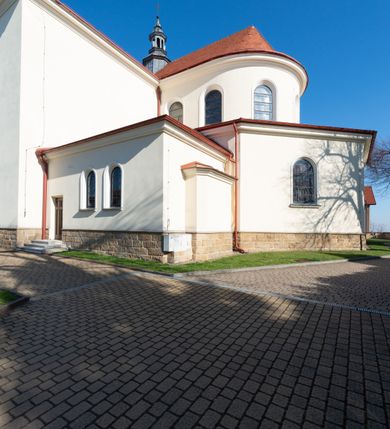 Zdjęcie nr 8: Kościół murowany, orientowany, jednonawowy, z transeptem, prezbiterium z obejściem i przybudówkami. Od zachodu kruchta, od północy kwadratowa kaplica, od południa kolejna przybudówka. 
Prezbiterium zamknięte półokrągło, z obejściem przeprutym do niego arkadami wspartymi na filarach. Ściany nawy i prezbiterium gładkie, przekryte sklepieniem kolebkowym z lunetami, w których mieszczą się okna. Transept i przecięcie naw sklepione krzyżowo. Na ścianach polichromia figuralna ze scenami z Nowego Testamentu, w partii sklepienia drobne, złote gwiazdki. Od zachodu chór zatopiony w występującym asymetrycznie murze, z tralkową balustradą. W prezbiterium siedem okien w kształcie stojącego prostokąta zamkniętego łukiem pełnym wypełnionych witrażami, w nawie sześć okien, w ramionach transeptu po jednym, czterodzielnym, wypełnionym witrażem, na wysokości chóru po dwa z każdej strony. Główne wejście od zachodu przez kruchtę. Fasada poprzedzona przedsionkiem o podziałach ramowych, z prostokątnym wejściem na osi, flankowanym parą pilastrów i prostokątnymi płycinami. Kondygnację wieńczy pełne belkowanie z wysuniętym gzymsem podtrzymującym daszek i trójkątny, schodkowy szczyt zakończony postumentami. Na centralnym krzyż, na sąsiadujących figury dzieci z rogami obfitości, a na zewnętrznych wazony płomieniste; szczyt ujmują postumenty z kulami. Fasada dwukondygnacyjna ze szczytem. Kondygnacja trójosiowa, rozdzielona pilastrami, o polach wypełnionych blendami w kształcie stojących prostokątów zamkniętych łukiem pełnym; szczyt trójkątny, z tarczą zegarową pośrodku. Budynek kościoła o wyraźnych podziałach na bryły składowe, ściany gładkie, przeprute oknami, z gzymsami pod krawędziami dachu. 
Nad nawą główną, na kalenicy, wieżyczka na sygnaturkę z latarnią i baniastym hełmem, druga na skrzyżowaniu naw, z latarnią i  hełmem zakończonym krzyżem. Dach nad nawą główną i przedsionkiem dwuspadowy, nad ramionami transeptu trójspadowym,nad prezbiterium zakończony wielobocznie, na przybudówkach dachy pulpitowe, dwuspadowe i wielospadowe. Dachy kryte dachówką ceramiczną, wieżyczki blachą. 
Najciekawszymi zabytkami kościoła są ołtarz główny z około 1700 roku z nastawą utworzoną z wici winnej latorośli, manierystyczny ołtarz boczny oraz siedemnastowieczna chrzcielnica z warsztatu Santi Gucciego. 
