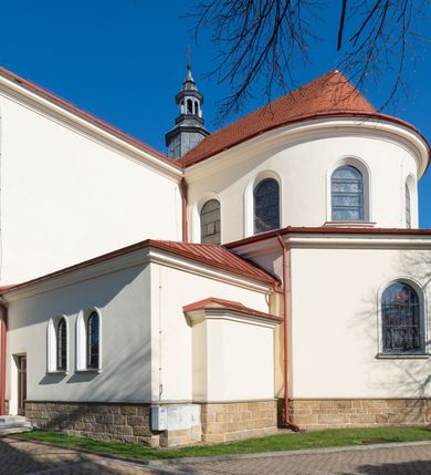 Zdjęcie nr 9: Kościół murowany, orientowany, jednonawowy, z transeptem, prezbiterium z obejściem i przybudówkami. Od zachodu kruchta, od północy kwadratowa kaplica, od południa kolejna przybudówka. 
Prezbiterium zamknięte półokrągło, z obejściem przeprutym do niego arkadami wspartymi na filarach. Ściany nawy i prezbiterium gładkie, przekryte sklepieniem kolebkowym z lunetami, w których mieszczą się okna. Transept i przecięcie naw sklepione krzyżowo. Na ścianach polichromia figuralna ze scenami z Nowego Testamentu, w partii sklepienia drobne, złote gwiazdki. Od zachodu chór zatopiony w występującym asymetrycznie murze, z tralkową balustradą. W prezbiterium siedem okien w kształcie stojącego prostokąta zamkniętego łukiem pełnym wypełnionych witrażami, w nawie sześć okien, w ramionach transeptu po jednym, czterodzielnym, wypełnionym witrażem, na wysokości chóru po dwa z każdej strony. Główne wejście od zachodu przez kruchtę. Fasada poprzedzona przedsionkiem o podziałach ramowych, z prostokątnym wejściem na osi, flankowanym parą pilastrów i prostokątnymi płycinami. Kondygnację wieńczy pełne belkowanie z wysuniętym gzymsem podtrzymującym daszek i trójkątny, schodkowy szczyt zakończony postumentami. Na centralnym krzyż, na sąsiadujących figury dzieci z rogami obfitości, a na zewnętrznych wazony płomieniste; szczyt ujmują postumenty z kulami. Fasada dwukondygnacyjna ze szczytem. Kondygnacja trójosiowa, rozdzielona pilastrami, o polach wypełnionych blendami w kształcie stojących prostokątów zamkniętych łukiem pełnym; szczyt trójkątny, z tarczą zegarową pośrodku. Budynek kościoła o wyraźnych podziałach na bryły składowe, ściany gładkie, przeprute oknami, z gzymsami pod krawędziami dachu. 
Nad nawą główną, na kalenicy, wieżyczka na sygnaturkę z latarnią i baniastym hełmem, druga na skrzyżowaniu naw, z latarnią i  hełmem zakończonym krzyżem. Dach nad nawą główną i przedsionkiem dwuspadowy, nad ramionami transeptu trójspadowym,nad prezbiterium zakończony wielobocznie, na przybudówkach dachy pulpitowe, dwuspadowe i wielospadowe. Dachy kryte dachówką ceramiczną, wieżyczki blachą. 
Najciekawszymi zabytkami kościoła są ołtarz główny z około 1700 roku z nastawą utworzoną z wici winnej latorośli, manierystyczny ołtarz boczny oraz siedemnastowieczna chrzcielnica z warsztatu Santi Gucciego. 
