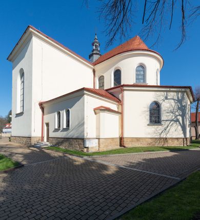 Zdjęcie nr 13: Kościół murowany, orientowany, jednonawowy, z transeptem, prezbiterium z obejściem i przybudówkami. Od zachodu kruchta, od północy kwadratowa kaplica, od południa kolejna przybudówka. 
Prezbiterium zamknięte półokrągło, z obejściem przeprutym do niego arkadami wspartymi na filarach. Ściany nawy i prezbiterium gładkie, przekryte sklepieniem kolebkowym z lunetami, w których mieszczą się okna. Transept i przecięcie naw sklepione krzyżowo. Na ścianach polichromia figuralna ze scenami z Nowego Testamentu, w partii sklepienia drobne, złote gwiazdki. Od zachodu chór zatopiony w występującym asymetrycznie murze, z tralkową balustradą. W prezbiterium siedem okien w kształcie stojącego prostokąta zamkniętego łukiem pełnym wypełnionych witrażami, w nawie sześć okien, w ramionach transeptu po jednym, czterodzielnym, wypełnionym witrażem, na wysokości chóru po dwa z każdej strony. Główne wejście od zachodu przez kruchtę. Fasada poprzedzona przedsionkiem o podziałach ramowych, z prostokątnym wejściem na osi, flankowanym parą pilastrów i prostokątnymi płycinami. Kondygnację wieńczy pełne belkowanie z wysuniętym gzymsem podtrzymującym daszek i trójkątny, schodkowy szczyt zakończony postumentami. Na centralnym krzyż, na sąsiadujących figury dzieci z rogami obfitości, a na zewnętrznych wazony płomieniste; szczyt ujmują postumenty z kulami. Fasada dwukondygnacyjna ze szczytem. Kondygnacja trójosiowa, rozdzielona pilastrami, o polach wypełnionych blendami w kształcie stojących prostokątów zamkniętych łukiem pełnym; szczyt trójkątny, z tarczą zegarową pośrodku. Budynek kościoła o wyraźnych podziałach na bryły składowe, ściany gładkie, przeprute oknami, z gzymsami pod krawędziami dachu. 
Nad nawą główną, na kalenicy, wieżyczka na sygnaturkę z latarnią i baniastym hełmem, druga na skrzyżowaniu naw, z latarnią i  hełmem zakończonym krzyżem. Dach nad nawą główną i przedsionkiem dwuspadowy, nad ramionami transeptu trójspadowym,nad prezbiterium zakończony wielobocznie, na przybudówkach dachy pulpitowe, dwuspadowe i wielospadowe. Dachy kryte dachówką ceramiczną, wieżyczki blachą. 
Najciekawszymi zabytkami kościoła są ołtarz główny z około 1700 roku z nastawą utworzoną z wici winnej latorośli, manierystyczny ołtarz boczny oraz siedemnastowieczna chrzcielnica z warsztatu Santi Gucciego. 
