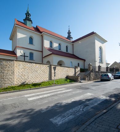 Zdjęcie nr 27: Kościół murowany, orientowany, jednonawowy, z transeptem, prezbiterium z obejściem i przybudówkami. Od zachodu kruchta, od północy kwadratowa kaplica, od południa kolejna przybudówka. 
Prezbiterium zamknięte półokrągło, z obejściem przeprutym do niego arkadami wspartymi na filarach. Ściany nawy i prezbiterium gładkie, przekryte sklepieniem kolebkowym z lunetami, w których mieszczą się okna. Transept i przecięcie naw sklepione krzyżowo. Na ścianach polichromia figuralna ze scenami z Nowego Testamentu, w partii sklepienia drobne, złote gwiazdki. Od zachodu chór zatopiony w występującym asymetrycznie murze, z tralkową balustradą. W prezbiterium siedem okien w kształcie stojącego prostokąta zamkniętego łukiem pełnym wypełnionych witrażami, w nawie sześć okien, w ramionach transeptu po jednym, czterodzielnym, wypełnionym witrażem, na wysokości chóru po dwa z każdej strony. Główne wejście od zachodu przez kruchtę. Fasada poprzedzona przedsionkiem o podziałach ramowych, z prostokątnym wejściem na osi, flankowanym parą pilastrów i prostokątnymi płycinami. Kondygnację wieńczy pełne belkowanie z wysuniętym gzymsem podtrzymującym daszek i trójkątny, schodkowy szczyt zakończony postumentami. Na centralnym krzyż, na sąsiadujących figury dzieci z rogami obfitości, a na zewnętrznych wazony płomieniste; szczyt ujmują postumenty z kulami. Fasada dwukondygnacyjna ze szczytem. Kondygnacja trójosiowa, rozdzielona pilastrami, o polach wypełnionych blendami w kształcie stojących prostokątów zamkniętych łukiem pełnym; szczyt trójkątny, z tarczą zegarową pośrodku. Budynek kościoła o wyraźnych podziałach na bryły składowe, ściany gładkie, przeprute oknami, z gzymsami pod krawędziami dachu. 
Nad nawą główną, na kalenicy, wieżyczka na sygnaturkę z latarnią i baniastym hełmem, druga na skrzyżowaniu naw, z latarnią i  hełmem zakończonym krzyżem. Dach nad nawą główną i przedsionkiem dwuspadowy, nad ramionami transeptu trójspadowym,nad prezbiterium zakończony wielobocznie, na przybudówkach dachy pulpitowe, dwuspadowe i wielospadowe. Dachy kryte dachówką ceramiczną, wieżyczki blachą. 
Najciekawszymi zabytkami kościoła są ołtarz główny z około 1700 roku z nastawą utworzoną z wici winnej latorośli, manierystyczny ołtarz boczny oraz siedemnastowieczna chrzcielnica z warsztatu Santi Gucciego. 
