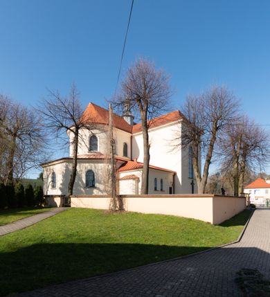 Zdjęcie nr 50: Kościół murowany, orientowany, jednonawowy, z transeptem, prezbiterium z obejściem i przybudówkami. Od zachodu kruchta, od północy kwadratowa kaplica, od południa kolejna przybudówka. 
Prezbiterium zamknięte półokrągło, z obejściem przeprutym do niego arkadami wspartymi na filarach. Ściany nawy i prezbiterium gładkie, przekryte sklepieniem kolebkowym z lunetami, w których mieszczą się okna. Transept i przecięcie naw sklepione krzyżowo. Na ścianach polichromia figuralna ze scenami z Nowego Testamentu, w partii sklepienia drobne, złote gwiazdki. Od zachodu chór zatopiony w występującym asymetrycznie murze, z tralkową balustradą. W prezbiterium siedem okien w kształcie stojącego prostokąta zamkniętego łukiem pełnym wypełnionych witrażami, w nawie sześć okien, w ramionach transeptu po jednym, czterodzielnym, wypełnionym witrażem, na wysokości chóru po dwa z każdej strony. Główne wejście od zachodu przez kruchtę. Fasada poprzedzona przedsionkiem o podziałach ramowych, z prostokątnym wejściem na osi, flankowanym parą pilastrów i prostokątnymi płycinami. Kondygnację wieńczy pełne belkowanie z wysuniętym gzymsem podtrzymującym daszek i trójkątny, schodkowy szczyt zakończony postumentami. Na centralnym krzyż, na sąsiadujących figury dzieci z rogami obfitości, a na zewnętrznych wazony płomieniste; szczyt ujmują postumenty z kulami. Fasada dwukondygnacyjna ze szczytem. Kondygnacja trójosiowa, rozdzielona pilastrami, o polach wypełnionych blendami w kształcie stojących prostokątów zamkniętych łukiem pełnym; szczyt trójkątny, z tarczą zegarową pośrodku. Budynek kościoła o wyraźnych podziałach na bryły składowe, ściany gładkie, przeprute oknami, z gzymsami pod krawędziami dachu. 
Nad nawą główną, na kalenicy, wieżyczka na sygnaturkę z latarnią i baniastym hełmem, druga na skrzyżowaniu naw, z latarnią i  hełmem zakończonym krzyżem. Dach nad nawą główną i przedsionkiem dwuspadowy, nad ramionami transeptu trójspadowym,nad prezbiterium zakończony wielobocznie, na przybudówkach dachy pulpitowe, dwuspadowe i wielospadowe. Dachy kryte dachówką ceramiczną, wieżyczki blachą. 
Najciekawszymi zabytkami kościoła są ołtarz główny z około 1700 roku z nastawą utworzoną z wici winnej latorośli, manierystyczny ołtarz boczny oraz siedemnastowieczna chrzcielnica z warsztatu Santi Gucciego. 
