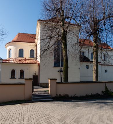 Zdjęcie nr 53: Kościół murowany, orientowany, jednonawowy, z transeptem, prezbiterium z obejściem i przybudówkami. Od zachodu kruchta, od północy kwadratowa kaplica, od południa kolejna przybudówka. 
Prezbiterium zamknięte półokrągło, z obejściem przeprutym do niego arkadami wspartymi na filarach. Ściany nawy i prezbiterium gładkie, przekryte sklepieniem kolebkowym z lunetami, w których mieszczą się okna. Transept i przecięcie naw sklepione krzyżowo. Na ścianach polichromia figuralna ze scenami z Nowego Testamentu, w partii sklepienia drobne, złote gwiazdki. Od zachodu chór zatopiony w występującym asymetrycznie murze, z tralkową balustradą. W prezbiterium siedem okien w kształcie stojącego prostokąta zamkniętego łukiem pełnym wypełnionych witrażami, w nawie sześć okien, w ramionach transeptu po jednym, czterodzielnym, wypełnionym witrażem, na wysokości chóru po dwa z każdej strony. Główne wejście od zachodu przez kruchtę. Fasada poprzedzona przedsionkiem o podziałach ramowych, z prostokątnym wejściem na osi, flankowanym parą pilastrów i prostokątnymi płycinami. Kondygnację wieńczy pełne belkowanie z wysuniętym gzymsem podtrzymującym daszek i trójkątny, schodkowy szczyt zakończony postumentami. Na centralnym krzyż, na sąsiadujących figury dzieci z rogami obfitości, a na zewnętrznych wazony płomieniste; szczyt ujmują postumenty z kulami. Fasada dwukondygnacyjna ze szczytem. Kondygnacja trójosiowa, rozdzielona pilastrami, o polach wypełnionych blendami w kształcie stojących prostokątów zamkniętych łukiem pełnym; szczyt trójkątny, z tarczą zegarową pośrodku. Budynek kościoła o wyraźnych podziałach na bryły składowe, ściany gładkie, przeprute oknami, z gzymsami pod krawędziami dachu. 
Nad nawą główną, na kalenicy, wieżyczka na sygnaturkę z latarnią i baniastym hełmem, druga na skrzyżowaniu naw, z latarnią i  hełmem zakończonym krzyżem. Dach nad nawą główną i przedsionkiem dwuspadowy, nad ramionami transeptu trójspadowym,nad prezbiterium zakończony wielobocznie, na przybudówkach dachy pulpitowe, dwuspadowe i wielospadowe. Dachy kryte dachówką ceramiczną, wieżyczki blachą. 
Najciekawszymi zabytkami kościoła są ołtarz główny z około 1700 roku z nastawą utworzoną z wici winnej latorośli, manierystyczny ołtarz boczny oraz siedemnastowieczna chrzcielnica z warsztatu Santi Gucciego. 
