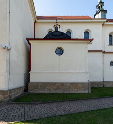 Zdjęcie nr 54: Kościół murowany, orientowany, jednonawowy, z transeptem, prezbiterium z obejściem i przybudówkami. Od zachodu kruchta, od północy kwadratowa kaplica, od południa kolejna przybudówka. 
Prezbiterium zamknięte półokrągło, z obejściem przeprutym do niego arkadami wspartymi na filarach. Ściany nawy i prezbiterium gładkie, przekryte sklepieniem kolebkowym z lunetami, w których mieszczą się okna. Transept i przecięcie naw sklepione krzyżowo. Na ścianach polichromia figuralna ze scenami z Nowego Testamentu, w partii sklepienia drobne, złote gwiazdki. Od zachodu chór zatopiony w występującym asymetrycznie murze, z tralkową balustradą. W prezbiterium siedem okien w kształcie stojącego prostokąta zamkniętego łukiem pełnym wypełnionych witrażami, w nawie sześć okien, w ramionach transeptu po jednym, czterodzielnym, wypełnionym witrażem, na wysokości chóru po dwa z każdej strony. Główne wejście od zachodu przez kruchtę. Fasada poprzedzona przedsionkiem o podziałach ramowych, z prostokątnym wejściem na osi, flankowanym parą pilastrów i prostokątnymi płycinami. Kondygnację wieńczy pełne belkowanie z wysuniętym gzymsem podtrzymującym daszek i trójkątny, schodkowy szczyt zakończony postumentami. Na centralnym krzyż, na sąsiadujących figury dzieci z rogami obfitości, a na zewnętrznych wazony płomieniste; szczyt ujmują postumenty z kulami. Fasada dwukondygnacyjna ze szczytem. Kondygnacja trójosiowa, rozdzielona pilastrami, o polach wypełnionych blendami w kształcie stojących prostokątów zamkniętych łukiem pełnym; szczyt trójkątny, z tarczą zegarową pośrodku. Budynek kościoła o wyraźnych podziałach na bryły składowe, ściany gładkie, przeprute oknami, z gzymsami pod krawędziami dachu. 
Nad nawą główną, na kalenicy, wieżyczka na sygnaturkę z latarnią i baniastym hełmem, druga na skrzyżowaniu naw, z latarnią i  hełmem zakończonym krzyżem. Dach nad nawą główną i przedsionkiem dwuspadowy, nad ramionami transeptu trójspadowym,nad prezbiterium zakończony wielobocznie, na przybudówkach dachy pulpitowe, dwuspadowe i wielospadowe. Dachy kryte dachówką ceramiczną, wieżyczki blachą. 
Najciekawszymi zabytkami kościoła są ołtarz główny z około 1700 roku z nastawą utworzoną z wici winnej latorośli, manierystyczny ołtarz boczny oraz siedemnastowieczna chrzcielnica z warsztatu Santi Gucciego. 
