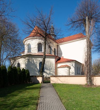 Zdjęcie nr 62: Kościół murowany, orientowany, jednonawowy, z transeptem, prezbiterium z obejściem i przybudówkami. Od zachodu kruchta, od północy kwadratowa kaplica, od południa kolejna przybudówka. 
Prezbiterium zamknięte półokrągło, z obejściem przeprutym do niego arkadami wspartymi na filarach. Ściany nawy i prezbiterium gładkie, przekryte sklepieniem kolebkowym z lunetami, w których mieszczą się okna. Transept i przecięcie naw sklepione krzyżowo. Na ścianach polichromia figuralna ze scenami z Nowego Testamentu, w partii sklepienia drobne, złote gwiazdki. Od zachodu chór zatopiony w występującym asymetrycznie murze, z tralkową balustradą. W prezbiterium siedem okien w kształcie stojącego prostokąta zamkniętego łukiem pełnym wypełnionych witrażami, w nawie sześć okien, w ramionach transeptu po jednym, czterodzielnym, wypełnionym witrażem, na wysokości chóru po dwa z każdej strony. Główne wejście od zachodu przez kruchtę. Fasada poprzedzona przedsionkiem o podziałach ramowych, z prostokątnym wejściem na osi, flankowanym parą pilastrów i prostokątnymi płycinami. Kondygnację wieńczy pełne belkowanie z wysuniętym gzymsem podtrzymującym daszek i trójkątny, schodkowy szczyt zakończony postumentami. Na centralnym krzyż, na sąsiadujących figury dzieci z rogami obfitości, a na zewnętrznych wazony płomieniste; szczyt ujmują postumenty z kulami. Fasada dwukondygnacyjna ze szczytem. Kondygnacja trójosiowa, rozdzielona pilastrami, o polach wypełnionych blendami w kształcie stojących prostokątów zamkniętych łukiem pełnym; szczyt trójkątny, z tarczą zegarową pośrodku. Budynek kościoła o wyraźnych podziałach na bryły składowe, ściany gładkie, przeprute oknami, z gzymsami pod krawędziami dachu. 
Nad nawą główną, na kalenicy, wieżyczka na sygnaturkę z latarnią i baniastym hełmem, druga na skrzyżowaniu naw, z latarnią i  hełmem zakończonym krzyżem. Dach nad nawą główną i przedsionkiem dwuspadowy, nad ramionami transeptu trójspadowym,nad prezbiterium zakończony wielobocznie, na przybudówkach dachy pulpitowe, dwuspadowe i wielospadowe. Dachy kryte dachówką ceramiczną, wieżyczki blachą. 
Najciekawszymi zabytkami kościoła są ołtarz główny z około 1700 roku z nastawą utworzoną z wici winnej latorośli, manierystyczny ołtarz boczny oraz siedemnastowieczna chrzcielnica z warsztatu Santi Gucciego. 
