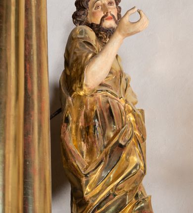 Zdjęcie nr 8: Rzeźba wydrążona, ścięta. Święty Jan Ewangelista ukazany w pozycji stojącej, w swobodnej pozie i znacznym kontrapoście, z lewą nogą ugiętą w kolanie. Jego prawa ręka jest zgięta w łokciu i uniesiona, w lewej przytrzymuje księgę. Głowa zwrócona w trzech czwartych, w prawo. Twarz okolona brodą, z rozchylonymi ustami. Na głowie włosy w postaci loków opadających na plecy. Święty jest ubrany w suknię i płaszcz zebrany w fałdach na lewym boku. Draperie o mocnych fałdowaniach, wyraźne, swobodnie rozwiana tkanina. Szaty i księga złocone, polichromia w partiach ciała naturalistyczna. 