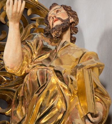 Zdjęcie nr 3: Rzeźba wydrążona, ścięta. Święty Jan Ewangelista ukazany w pozycji stojącej, w swobodnej pozie i znacznym kontrapoście, z lewą nogą ugiętą w kolanie. Jego prawa ręka jest zgięta w łokciu i uniesiona, w lewej przytrzymuje księgę. Głowa zwrócona w trzech czwartych, w prawo. Twarz okolona brodą, z rozchylonymi ustami. Na głowie włosy w postaci loków opadających na plecy. Święty jest ubrany w suknię i płaszcz zebrany w fałdach na lewym boku. Draperie o mocnych fałdowaniach, wyraźne, swobodnie rozwiana tkanina. Szaty i księga złocone, polichromia w partiach ciała naturalistyczna. 