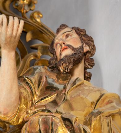 Zdjęcie nr 2: Rzeźba wydrążona, ścięta. Święty Jan Ewangelista ukazany w pozycji stojącej, w swobodnej pozie i znacznym kontrapoście, z lewą nogą ugiętą w kolanie. Jego prawa ręka jest zgięta w łokciu i uniesiona, w lewej przytrzymuje księgę. Głowa zwrócona w trzech czwartych, w prawo. Twarz okolona brodą, z rozchylonymi ustami. Na głowie włosy w postaci loków opadających na plecy. Święty jest ubrany w suknię i płaszcz zebrany w fałdach na lewym boku. Draperie o mocnych fałdowaniach, wyraźne, swobodnie rozwiana tkanina. Szaty i księga złocone, polichromia w partiach ciała naturalistyczna. 