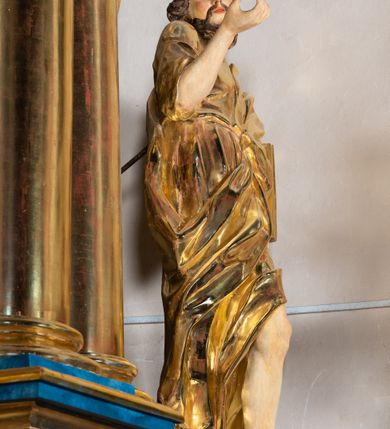 Zdjęcie nr 7: Rzeźba wydrążona, ścięta. Święty Jan Ewangelista ukazany w pozycji stojącej, w swobodnej pozie i znacznym kontrapoście, z lewą nogą ugiętą w kolanie. Jego prawa ręka jest zgięta w łokciu i uniesiona, w lewej przytrzymuje księgę. Głowa zwrócona w trzech czwartych, w prawo. Twarz okolona brodą, z rozchylonymi ustami. Na głowie włosy w postaci loków opadających na plecy. Święty jest ubrany w suknię i płaszcz zebrany w fałdach na lewym boku. Draperie o mocnych fałdowaniach, wyraźne, swobodnie rozwiana tkanina. Szaty i księga złocone, polichromia w partiach ciała naturalistyczna. 