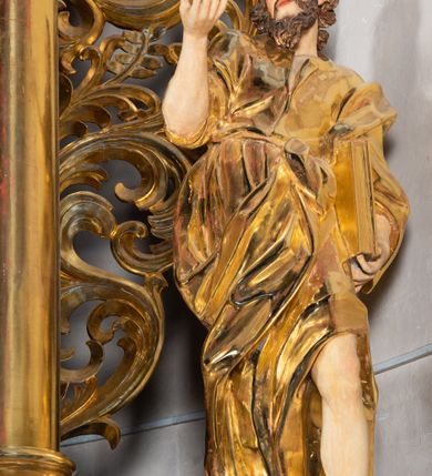 Zdjęcie nr 9: Rzeźba wydrążona, ścięta. Święty Jan Ewangelista ukazany w pozycji stojącej, w swobodnej pozie i znacznym kontrapoście, z lewą nogą ugiętą w kolanie. Jego prawa ręka jest zgięta w łokciu i uniesiona, w lewej przytrzymuje księgę. Głowa zwrócona w trzech czwartych, w prawo. Twarz okolona brodą, z rozchylonymi ustami. Na głowie włosy w postaci loków opadających na plecy. Święty jest ubrany w suknię i płaszcz zebrany w fałdach na lewym boku. Draperie o mocnych fałdowaniach, wyraźne, swobodnie rozwiana tkanina. Szaty i księga złocone, polichromia w partiach ciała naturalistyczna. 