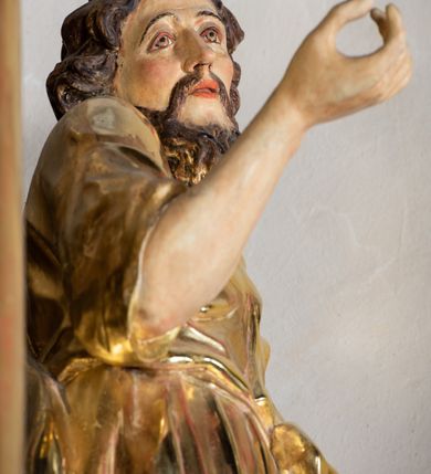 Zdjęcie nr 5: Rzeźba wydrążona, ścięta. Święty Jan Ewangelista ukazany w pozycji stojącej, w swobodnej pozie i znacznym kontrapoście, z lewą nogą ugiętą w kolanie. Jego prawa ręka jest zgięta w łokciu i uniesiona, w lewej przytrzymuje księgę. Głowa zwrócona w trzech czwartych, w prawo. Twarz okolona brodą, z rozchylonymi ustami. Na głowie włosy w postaci loków opadających na plecy. Święty jest ubrany w suknię i płaszcz zebrany w fałdach na lewym boku. Draperie o mocnych fałdowaniach, wyraźne, swobodnie rozwiana tkanina. Szaty i księga złocone, polichromia w partiach ciała naturalistyczna. 