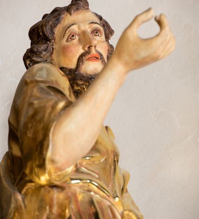 Zdjęcie nr 6: Rzeźba wydrążona, ścięta. Święty Jan Ewangelista ukazany w pozycji stojącej, w swobodnej pozie i znacznym kontrapoście, z lewą nogą ugiętą w kolanie. Jego prawa ręka jest zgięta w łokciu i uniesiona, w lewej przytrzymuje księgę. Głowa zwrócona w trzech czwartych, w prawo. Twarz okolona brodą, z rozchylonymi ustami. Na głowie włosy w postaci loków opadających na plecy. Święty jest ubrany w suknię i płaszcz zebrany w fałdach na lewym boku. Draperie o mocnych fałdowaniach, wyraźne, swobodnie rozwiana tkanina. Szaty i księga złocone, polichromia w partiach ciała naturalistyczna. 