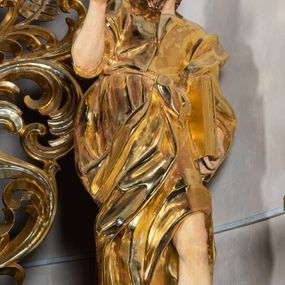 Zdjęcie nr 1: Rzeźba wydrążona, ścięta. Święty Jan Ewangelista ukazany w pozycji stojącej, w swobodnej pozie i znacznym kontrapoście, z lewą nogą ugiętą w kolanie. Jego prawa ręka jest zgięta w łokciu i uniesiona, w lewej przytrzymuje księgę. Głowa zwrócona w trzech czwartych, w prawo. Twarz okolona brodą, z rozchylonymi ustami. Na głowie włosy w postaci loków opadających na plecy. Święty jest ubrany w suknię i płaszcz zebrany w fałdach na lewym boku. Draperie o mocnych fałdowaniach, wyraźne, swobodnie rozwiana tkanina. Szaty i księga złocone, polichromia w partiach ciała naturalistyczna. 