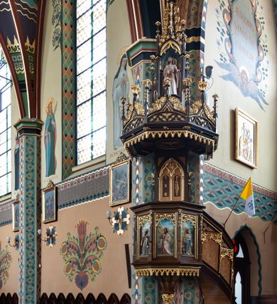 Zdjęcie nr 1: Ambona neogotycka, zawieszona na narożnym filarze nawy głównej i północnego ramienia transeptu. Na ambonę prowadzą schody z lekko skręconą balustradą przylegającą do północnej strony prezbiterium. Balustrada artykułowana romboidalnymi płycinami podzielonymi na pola w kształcie trapezu i małego trójkąta. W polach obu płycin białe dekoracje ze stylizowanej wici akantu; płyciny trapezowe u góry zdobione rzeźbioną, ażurową dekoracją stylizowanego astwerku. Kosz na planie sześcioboku ujęty od góry i od dołu profilowanym gzymsem. Podstawa ośmioboczna, zwężająca się ku dołowi, ujęta pozłacanym i ażurowym ornamentem roślinnym. Boki kosza z czterema płycinami, flankowanymi wąskimi filarkami ze szkarpami i zwieńczonymi pinaklami. W polach płycin ukazane są płaskorzeźbione wizerunki ewangelistów, od lewej strony: św. Mateusza, św. Jana, św. Łukasza i św. Marka. Ewangeliści przedstawieni w postawie stojącej z księgami i piórami do pisania w dłoniach. U ich stóp znajdują się odpowiadające im atrybuty. Sylwetki polichromowane, ukazane na tle malowanego pejzażu. Płyciny zwieńczone rzeźbioną, ażurową dekoracją astwerku. Zaplecek prostokątny ujęty dwoma sterczynami wspartymi na niewielkich konsolkach. W polu zaplecka ostrołukowa arkada z polem przedzielonym maswerkową dekoracją wspartą na trzech półkolumienkach. W polu dwie tablice Dekalogu. Baldachim ambony rozłożysty, powtarzający plan kosza. Podniebie podzielone promieniście odchodzącymi listwami, pośrodku ośmioboczne pole z płaskorzeźbioną gołębicą Ducha Świętego. W zwieńczeniu architektoniczna aedicula wsparta na czterech kolumnach z głęboką wnęką, w której umieszczona jest rzeźba Chrystusa. Aedicula zwieńczona jest wysoką sterczyną zakończoną łacińskim krzyżem. Baldachim zamknięty fryzem wypełnionym astwerkiem i ażurowym ornamentem z wolutowo skręconej wici roślinnej. W narożach sterczyny z żabkami i kwiatonami. Spód kosza i baldachimu zdobione są rzeźbionymi arkadkami. Struktura ambony malowana w kolorze ciemnobrązowym, płyciny i balustrady jasnobrązowe, detale architektoniczne i ornamentyka złocone.

