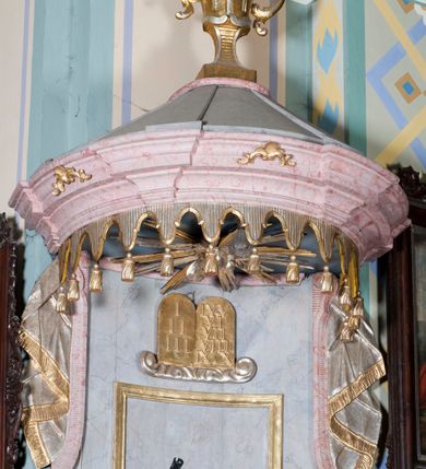 Zdjęcie nr 3: Ambona, przyścienna, wisząca, z jednobiegowymi schodami od strony prezbiterium. Balustrada dekorowana romboidalną płyciną z dekoracją z ornamentu rocaille’owego i akantowego. Poręcz w formie profilowanego gzymsu. Kosz na planie odcinka koła, ściany kosza wypukłe, podzielone wolutami, w polach płyciny wypełnione dekoracją w formie zbliżonej do owalu utworzonego z ornamentu rocaille’owego. Parapet w kształcie profilowanego gzymsu, wyłamanego na osi wolut. Kosz od dołu zakończony stożkiem ujętym czterema wolutami w formie karbowanej wstęgi, dekorowany motywem roślinnym, zakończony szyszką. Zaplecek wklęsły, flankowany spływami wolutowymi oraz udrapowaną tkaniną. W polu prostokątna płycina, powyżej płaskorzeźbione tablice mojżeszowe na obłoku. Baldachim powielający plan kosza, z wyłamującym się gzymsem na osiach, z podwieszonym lambrekinem, zwężający się ku górze, zwieńczony rokokowym wazonem z płomieniem. W podniebiu baldachimu rzeźbiona gołębica Ducha Świętego w glorii promienistej. Struktura marmoryzowana w kolorze jasnoniebieskim, profile w jasnoróżowym, dekoracje złocone i srebrzone.