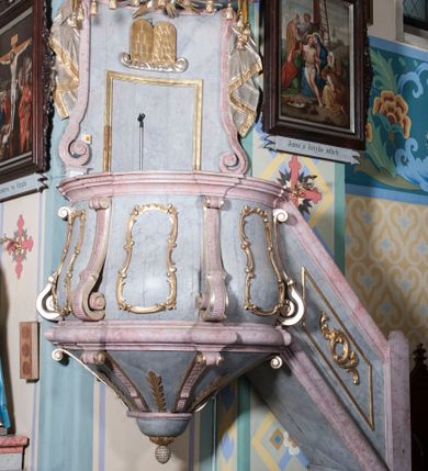 Zdjęcie nr 1: Ambona, przyścienna, wisząca, z jednobiegowymi schodami od strony prezbiterium. Balustrada dekorowana romboidalną płyciną z dekoracją z ornamentu rocaille’owego i akantowego. Poręcz w formie profilowanego gzymsu. Kosz na planie odcinka koła, ściany kosza wypukłe, podzielone wolutami, w polach płyciny wypełnione dekoracją w formie zbliżonej do owalu utworzonego z ornamentu rocaille’owego. Parapet w kształcie profilowanego gzymsu, wyłamanego na osi wolut. Kosz od dołu zakończony stożkiem ujętym czterema wolutami w formie karbowanej wstęgi, dekorowany motywem roślinnym, zakończony szyszką. Zaplecek wklęsły, flankowany spływami wolutowymi oraz udrapowaną tkaniną. W polu prostokątna płycina, powyżej płaskorzeźbione tablice mojżeszowe na obłoku. Baldachim powielający plan kosza, z wyłamującym się gzymsem na osiach, z podwieszonym lambrekinem, zwężający się ku górze, zwieńczony rokokowym wazonem z płomieniem. W podniebiu baldachimu rzeźbiona gołębica Ducha Świętego w glorii promienistej. Struktura marmoryzowana w kolorze jasnoniebieskim, profile w jasnoróżowym, dekoracje złocone i srebrzone.