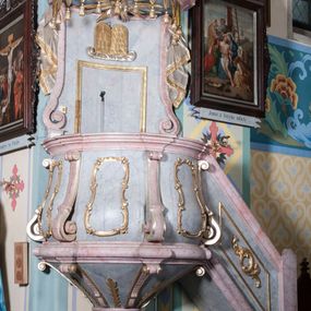 Zdjęcie nr 1: Ambona, przyścienna, wisząca, z jednobiegowymi schodami od strony prezbiterium. Balustrada dekorowana romboidalną płyciną z dekoracją z ornamentu rocaille’owego i akantowego. Poręcz w formie profilowanego gzymsu. Kosz na planie odcinka koła, ściany kosza wypukłe, podzielone wolutami, w polach płyciny wypełnione dekoracją w formie zbliżonej do owalu utworzonego z ornamentu rocaille’owego. Parapet w kształcie profilowanego gzymsu, wyłamanego na osi wolut. Kosz od dołu zakończony stożkiem ujętym czterema wolutami w formie karbowanej wstęgi, dekorowany motywem roślinnym, zakończony szyszką. Zaplecek wklęsły, flankowany spływami wolutowymi oraz udrapowaną tkaniną. W polu prostokątna płycina, powyżej płaskorzeźbione tablice mojżeszowe na obłoku. Baldachim powielający plan kosza, z wyłamującym się gzymsem na osiach, z podwieszonym lambrekinem, zwężający się ku górze, zwieńczony rokokowym wazonem z płomieniem. W podniebiu baldachimu rzeźbiona gołębica Ducha Świętego w glorii promienistej. Struktura marmoryzowana w kolorze jasnoniebieskim, profile w jasnoróżowym, dekoracje złocone i srebrzone.