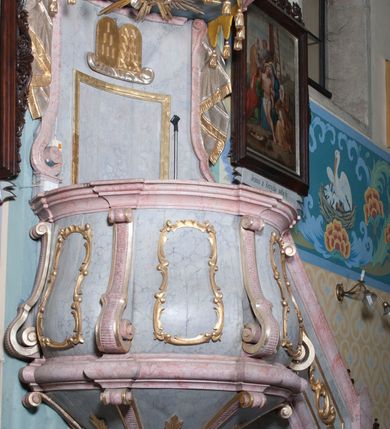 Zdjęcie nr 2: Ambona, przyścienna, wisząca, z jednobiegowymi schodami od strony prezbiterium. Balustrada dekorowana romboidalną płyciną z dekoracją z ornamentu rocaille’owego i akantowego. Poręcz w formie profilowanego gzymsu. Kosz na planie odcinka koła, ściany kosza wypukłe, podzielone wolutami, w polach płyciny wypełnione dekoracją w formie zbliżonej do owalu utworzonego z ornamentu rocaille’owego. Parapet w kształcie profilowanego gzymsu, wyłamanego na osi wolut. Kosz od dołu zakończony stożkiem ujętym czterema wolutami w formie karbowanej wstęgi, dekorowany motywem roślinnym, zakończony szyszką. Zaplecek wklęsły, flankowany spływami wolutowymi oraz udrapowaną tkaniną. W polu prostokątna płycina, powyżej płaskorzeźbione tablice mojżeszowe na obłoku. Baldachim powielający plan kosza, z wyłamującym się gzymsem na osiach, z podwieszonym lambrekinem, zwężający się ku górze, zwieńczony rokokowym wazonem z płomieniem. W podniebiu baldachimu rzeźbiona gołębica Ducha Świętego w glorii promienistej. Struktura marmoryzowana w kolorze jasnoniebieskim, profile w jasnoróżowym, dekoracje złocone i srebrzone.