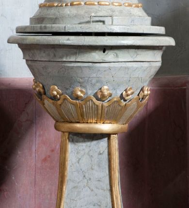 Zdjęcie nr 1: Antyambona złączona z chrzcielnicą. Stopa chrzcielnicy na planie kwadratu, profilowana. Trzon czteroboczny, zwężający się ku górze, w narożach od frontu ujęty listwami. Czara w formie odwróconego, ściętego stożka, z wywiniętą krawędzią. Od spodu opięta pierścieniem i silnie uproszczonym, stylizowanym, żłobkowanym lambrekinem. Pokrywa w formie profilowanego, ściętego stożka, zwieńczonego akantem i szyszką. We wnętrzu czary miedziana misa oraz fragment drewnianej pokrywy z rozetą. Antyambona przyścienna, o wklęsłym licu, na cokole, ujęta filarami podtrzymującymi profilowany gzyms, dekorowanymi płycinami. Pośrodku wspornik w formie wybrzuszonego stożka, o ściętym wierzchołku z grupą rzeźbiarską Chrzest Chrystusa. Powyżej zaplecek ujęty pilastrami, dźwigającymi spływ wolutowe; po bokach udrapowana tkanina. W polu środkowym płycina z rzeźbioną gołębicą Ducha Świętego na tle glorii promienistej. Baldachim na planie odcinka koła, z wyłamującym się gzymsem, dekorowany podwieszonym lambrekinem, zwężający się ku górze, zakończony rokokowym wazonem płomienistym. Struktura marmoryzowana w kolorze jasnoniebieskim, cokół, gzyms i profile jasnoróżowe, dekoracja złocona i srebrzona.