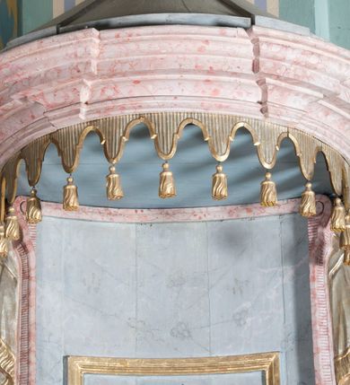 Zdjęcie nr 3: Antyambona złączona z chrzcielnicą. Stopa chrzcielnicy na planie kwadratu, profilowana. Trzon czteroboczny, zwężający się ku górze, w narożach od frontu ujęty listwami. Czara w formie odwróconego, ściętego stożka, z wywiniętą krawędzią. Od spodu opięta pierścieniem i silnie uproszczonym, stylizowanym, żłobkowanym lambrekinem. Pokrywa w formie profilowanego, ściętego stożka, zwieńczonego akantem i szyszką. We wnętrzu czary miedziana misa oraz fragment drewnianej pokrywy z rozetą. Antyambona przyścienna, o wklęsłym licu, na cokole, ujęta filarami podtrzymującymi profilowany gzyms, dekorowanymi płycinami. Pośrodku wspornik w formie wybrzuszonego stożka, o ściętym wierzchołku z grupą rzeźbiarską Chrzest Chrystusa. Powyżej zaplecek ujęty pilastrami, dźwigającymi spływ wolutowe; po bokach udrapowana tkanina. W polu środkowym płycina z rzeźbioną gołębicą Ducha Świętego na tle glorii promienistej. Baldachim na planie odcinka koła, z wyłamującym się gzymsem, dekorowany podwieszonym lambrekinem, zwężający się ku górze, zakończony rokokowym wazonem płomienistym. Struktura marmoryzowana w kolorze jasnoniebieskim, cokół, gzyms i profile jasnoróżowe, dekoracja złocona i srebrzona.