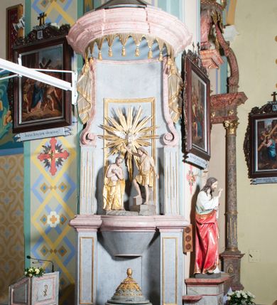 Zdjęcie nr 7: Antyambona złączona z chrzcielnicą. Stopa chrzcielnicy na planie kwadratu, profilowana. Trzon czteroboczny, zwężający się ku górze, w narożach od frontu ujęty listwami. Czara w formie odwróconego, ściętego stożka, z wywiniętą krawędzią. Od spodu opięta pierścieniem i silnie uproszczonym, stylizowanym, żłobkowanym lambrekinem. Pokrywa w formie profilowanego, ściętego stożka, zwieńczonego akantem i szyszką. We wnętrzu czary miedziana misa oraz fragment drewnianej pokrywy z rozetą. Antyambona przyścienna, o wklęsłym licu, na cokole, ujęta filarami podtrzymującymi profilowany gzyms, dekorowanymi płycinami. Pośrodku wspornik w formie wybrzuszonego stożka, o ściętym wierzchołku z grupą rzeźbiarską Chrzest Chrystusa. Powyżej zaplecek ujęty pilastrami, dźwigającymi spływ wolutowe; po bokach udrapowana tkanina. W polu środkowym płycina z rzeźbioną gołębicą Ducha Świętego na tle glorii promienistej. Baldachim na planie odcinka koła, z wyłamującym się gzymsem, dekorowany podwieszonym lambrekinem, zwężający się ku górze, zakończony rokokowym wazonem płomienistym. Struktura marmoryzowana w kolorze jasnoniebieskim, cokół, gzyms i profile jasnoróżowe, dekoracja złocona i srebrzona.