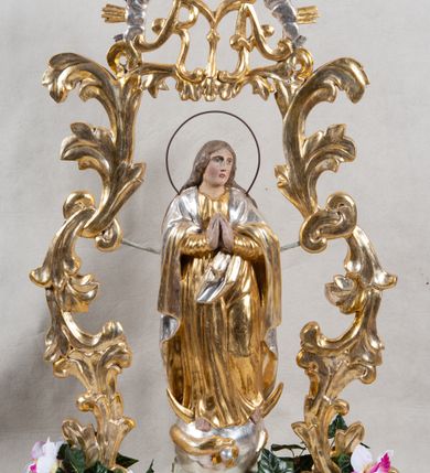 Zdjęcie nr 2: Feretron na profilowanej, prostopadłościennej podstawie z przedstawieniem Maki Boskiej Niepokalanej. Rzeźba pełna została ustawiona na profilowanym cokole, półksiężycu i globie, otoczonym przez wijącego się węża z owocem (jabłkiem) w paszczy. Maria została ukazana frontalnie, w kontrapoście, z dłońmi złożonymi w geście modlitwy, z głową przechyloną na lewe ramię. Twarz ma owalną, o pełnych policzkach i głęboko osadzonych oczach; włosy jasne, długie, opadające na plecy i ramiona. Maria jest ubrana w złotą suknię, o wąskich rękawach i narzucony na ramiona srebrny płaszcz. Figura została ujęta rzeźbionymi liśćmi akantu podtrzymującymi monogram „MARYA” w otoczeniu obłoków i glorii. Polichromia naturalistyczna w odsłoniętych partiach ciała; szaty i dekoracja ornamentalna złocona i srebrzona.