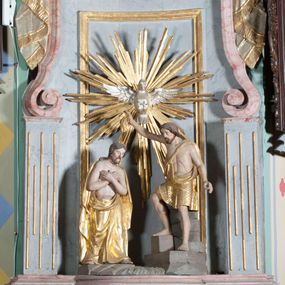 Zdjęcie nr 1: Grupa rzeźbiarska przedstawiająca scenę Chrztu Chrystusa. Rzeźby pełne, całopostaciowe. Po prawej stronie, na blikach skalnych św. Jan Chrzciciel zwrócony w prawo, z prawą nogą ustawioną wyżej, z uniesioną prawą ręką, w której trzyma muszlę z wodą, którą wylewa nad głową Chrystusa. Twarz ma pociągłą, szczupłą, okoloną krótką brodą, włosy półdługie, opadające na kark. Ubrany w osłaniający biodra płaszcz ze skóry, podtrzymywany przez taśmę przewiązaną przez lewe ramię. Po przeciwnej stronie figura Chrystusa zwróconego w trzech czwartych w lewo, w kontrapoście, delikatnie pochylonego z dłońmi skrzyżowanymi na piersi. Twarz pociągła, okolona krótką brodą, włosy długie, opadające na plecy. Na ramiona ma zarzucony płaszcz, biodra przepasane drapowaną tkaniną sięgającą kostek, tors nagi. W tle dużej wielkości gołębica Ducha Świętego na tle glorii promienistej. W odsłoniętych partiach ciała polichromia naturalistyczna, szaty złocone. 
