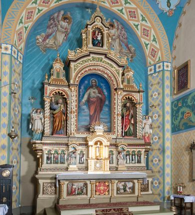 Zdjęcie nr 3: Nastawa ołtarzowa jednokondygnacyjna ze zwieńczeniem, trójosiowa, na cokole. Mensa prostopadłościenna, murowana, w drewnianej obudowie, z kamiennym blatem. Antepedium podzielone na trzy pola, wydzielone czterema kolumienkami. Zewnętrzne płyciny w formie leżącego prostokąta, z płaskorzeźbionymi przedstawieniami po lewej stronie ofiara Melchizedeka, po prawej ofiara Abrahama. W polu środkowym płycina w formie kwadratu, z stylizowanym maswerkowaniem i równoramiennym krzyżem pośrodku oraz rozetami w narożach. Na mensie tabernakulum na wysokiej, prostopadłościennej podstawie, w której znajdują się drzwiczki dekorowane hostią w glorii, flankowane pilastrami z dekoracją  ornamentem arabeskowym. Na osi tron wystawienia w formie niszy ujęty parą smukłych kolumienek podtrzymujących szerokie belkowanie przechodzące nad niszą w łuk. Gzyms wieńczy ornament roślinny, na środku na cokole postać baranka w glorii na księdze z siedmioma pieczęciami. Predella podzielona na trzy pola z występującymi do przodu dwiema niszami z figurami klęczących aniołów. W polach zewnętrznych trzy arkadki z płaskorzeźbionymi przedstawieniami świętych. W polu środkowym tabernakulum flankowane przed dwie arkadki z przedstawieniami świętych. Pierwsza kondygnacja utworzona z trzech arkad wydzielonych kolumnami, oś środkowa wyższa od bocznych. W polu środkowym obraz z przedstawieniem Najświętszego Serca Jezusa, w profilowanej ramie; archiwolta ujęta kolumienkami. Całość zamknięta silnie wysuniętym gzymsem o fantazyjnej linii. W osiach bocznych aedicule z posągami św. Piotra i św. Pawła, zwieńczone daszkiem z latarniami. Kondygnacja flankowana wielobocznymi wspornikami z figurami aniołów. Zwieńczenie w formie aediculi ujętej kolumienkami we wnękach oraz otwartymi półarkadami. Całość zamknięta gzymsem o fantazyjnej linii oraz krzyżem. W niszy figura Matki Boskiej z Dzieciątkiem. Cokoły zdobione płycinami z malowaną i złoconą dekoracja roślinną. Rama obrazu w polu środkowym dekorowana ornamentem roślinno-geometrycznym; archiwolta ornamentem roślinnym w formie liści. Powyżej płycina w kształcie łuku odcinkowego z rzeźbionym ornamentem roślinnym. Archiwolty arkad na osiach bocznych dekorowane ornamentem geometrycznym i stylizowanymi żabkami, w przyłuczach rozety; latarnie zdobione ornamentem roślinnym i stylizowanymi żabkami. Struktura polichromowana w kolorze kości słoniowej i szarym; płyciny i profilowania w kolorze czerwonym oraz zielonym, elementy dekoracyjne złocone.