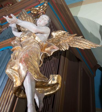 Zdjęcie nr 2: Rzeźby drążone, ścięte z tyłu. Figury aniołów zwrócone frontalnie, unoszące się z ugiętymi swobodnie nogami; o rozpostartych skrzydłach. Anioł po lewej stronie z wniesionymi do góry rękami. Ma podłużną, pełną twarz o młodzieńczych rysach, półdługich, kręconych włosach. Biodra przepasane sięgającą łydek, drapowaną, ekspresyjnie rozwianą tkaniną, podwiązaną na lewym ramieniu wstążką. Anioł z prawej strony z głową przechyloną na lewe ramię, prawą ręką uniesioną do góry i lewą wyciągniętą przed siebie. Ma podobną twarz o na wpół przymkniętych oczach; włosy półdługie, falowane. Ubrany w szatę odsłaniającą lewe ramię, przepasaną w talii oraz płaszcz opasujący biodra. Polichromia naturalistyczna w odsłoniętych partiach ciała, szaty i skrzydła złocone.