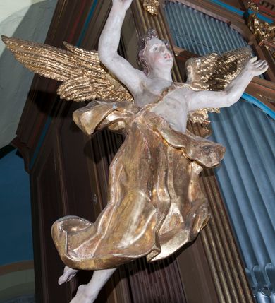 Zdjęcie nr 1: Rzeźby drążone, ścięte z tyłu. Figury aniołów zwrócone frontalnie, unoszące się z ugiętymi swobodnie nogami; o rozpostartych skrzydłach. Anioł po lewej stronie z wniesionymi do góry rękami. Ma podłużną, pełną twarz o młodzieńczych rysach, półdługich, kręconych włosach. Biodra przepasane sięgającą łydek, drapowaną, ekspresyjnie rozwianą tkaniną, podwiązaną na lewym ramieniu wstążką. Anioł z prawej strony z głową przechyloną na lewe ramię, prawą ręką uniesioną do góry i lewą wyciągniętą przed siebie. Ma podobną twarz o na wpół przymkniętych oczach; włosy półdługie, falowane. Ubrany w szatę odsłaniającą lewe ramię, przepasaną w talii oraz płaszcz opasujący biodra. Polichromia naturalistyczna w odsłoniętych partiach ciała, szaty i skrzydła złocone.