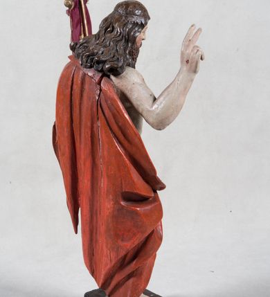 Zdjęcie nr 5: Rzeźba pełna ukazująca Chrystusa Zmartwychwstałego przedstawionego całopostaciowo, w lekkim kontrapoście, z delikatnie zaznaczoną muskulaturą i głową lekko opuszczoną. Jezus ma jasną karnację, twarz pociągłą o prostym nosie, okoloną długą brodą i falującymi brązowymi włosami spływającymi na plecy i prawe ramię. Jest ubrany w udrapowany czerwony płaszcz w diagonalnym układzie zarzucony na lewe ramię i owijający się wokół prawego biodra, łączący się pod lewą ręką. W lewej dłoni wtórnie dodana chorągiew w postaci purpurowej tkaniny z krzyżem na drewnianym drzewcu z resztkami złocenia, prawa dłoń wzniesiona w geście błogosławieństwa. Postać ustawiona na niewysokim postumencie imitującym skalny blok. 