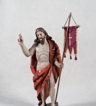 Zdjęcie nr 1: Rzeźba pełna ukazująca Chrystusa Zmartwychwstałego przedstawionego całopostaciowo, w lekkim kontrapoście, z delikatnie zaznaczoną muskulaturą i głową lekko opuszczoną. Jezus ma jasną karnację, twarz pociągłą o prostym nosie, okoloną długą brodą i falującymi brązowymi włosami spływającymi na plecy i prawe ramię. Jest ubrany w udrapowany czerwony płaszcz w diagonalnym układzie zarzucony na lewe ramię i owijający się wokół prawego biodra, łączący się pod lewą ręką. W lewej dłoni wtórnie dodana chorągiew w postaci purpurowej tkaniny z krzyżem na drewnianym drzewcu z resztkami złocenia, prawa dłoń wzniesiona w geście błogosławieństwa. Postać ustawiona na niewysokim postumencie imitującym skalny blok. 