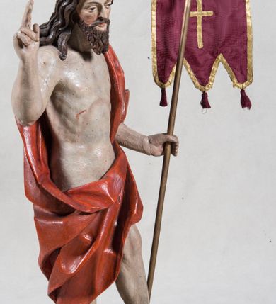 Zdjęcie nr 2: Rzeźba pełna ukazująca Chrystusa Zmartwychwstałego przedstawionego całopostaciowo, w lekkim kontrapoście, z delikatnie zaznaczoną muskulaturą i głową lekko opuszczoną. Jezus ma jasną karnację, twarz pociągłą o prostym nosie, okoloną długą brodą i falującymi brązowymi włosami spływającymi na plecy i prawe ramię. Jest ubrany w udrapowany czerwony płaszcz w diagonalnym układzie zarzucony na lewe ramię i owijający się wokół prawego biodra, łączący się pod lewą ręką. W lewej dłoni wtórnie dodana chorągiew w postaci purpurowej tkaniny z krzyżem na drewnianym drzewcu z resztkami złocenia, prawa dłoń wzniesiona w geście błogosławieństwa. Postać ustawiona na niewysokim postumencie imitującym skalny blok. 