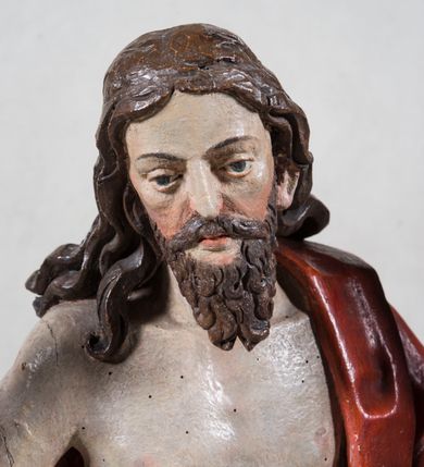 Zdjęcie nr 8: Rzeźba pełna ukazująca Chrystusa Zmartwychwstałego przedstawionego całopostaciowo, w lekkim kontrapoście, z delikatnie zaznaczoną muskulaturą i głową lekko opuszczoną. Jezus ma jasną karnację, twarz pociągłą o prostym nosie, okoloną długą brodą i falującymi brązowymi włosami spływającymi na plecy i prawe ramię. Jest ubrany w udrapowany czerwony płaszcz w diagonalnym układzie zarzucony na lewe ramię i owijający się wokół prawego biodra, łączący się pod lewą ręką. W lewej dłoni wtórnie dodana chorągiew w postaci purpurowej tkaniny z krzyżem na drewnianym drzewcu z resztkami złocenia, prawa dłoń wzniesiona w geście błogosławieństwa. Postać ustawiona na niewysokim postumencie imitującym skalny blok. 