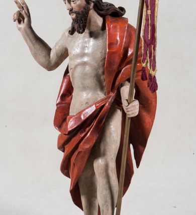 Zdjęcie nr 3: Rzeźba pełna ukazująca Chrystusa Zmartwychwstałego przedstawionego całopostaciowo, w lekkim kontrapoście, z delikatnie zaznaczoną muskulaturą i głową lekko opuszczoną. Jezus ma jasną karnację, twarz pociągłą o prostym nosie, okoloną długą brodą i falującymi brązowymi włosami spływającymi na plecy i prawe ramię. Jest ubrany w udrapowany czerwony płaszcz w diagonalnym układzie zarzucony na lewe ramię i owijający się wokół prawego biodra, łączący się pod lewą ręką. W lewej dłoni wtórnie dodana chorągiew w postaci purpurowej tkaniny z krzyżem na drewnianym drzewcu z resztkami złocenia, prawa dłoń wzniesiona w geście błogosławieństwa. Postać ustawiona na niewysokim postumencie imitującym skalny blok. 