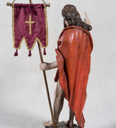 Zdjęcie nr 7: Rzeźba pełna ukazująca Chrystusa Zmartwychwstałego przedstawionego całopostaciowo, w lekkim kontrapoście, z delikatnie zaznaczoną muskulaturą i głową lekko opuszczoną. Jezus ma jasną karnację, twarz pociągłą o prostym nosie, okoloną długą brodą i falującymi brązowymi włosami spływającymi na plecy i prawe ramię. Jest ubrany w udrapowany czerwony płaszcz w diagonalnym układzie zarzucony na lewe ramię i owijający się wokół prawego biodra, łączący się pod lewą ręką. W lewej dłoni wtórnie dodana chorągiew w postaci purpurowej tkaniny z krzyżem na drewnianym drzewcu z resztkami złocenia, prawa dłoń wzniesiona w geście błogosławieństwa. Postać ustawiona na niewysokim postumencie imitującym skalny blok. 