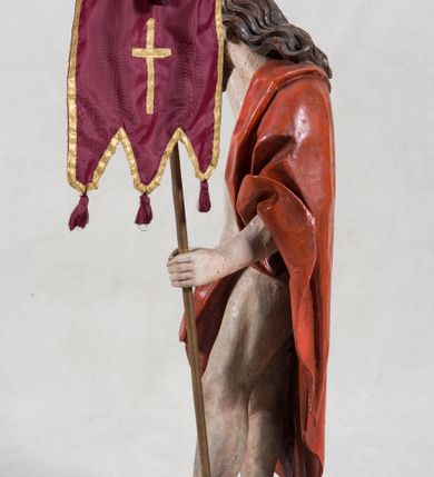 Zdjęcie nr 4: Rzeźba pełna ukazująca Chrystusa Zmartwychwstałego przedstawionego całopostaciowo, w lekkim kontrapoście, z delikatnie zaznaczoną muskulaturą i głową lekko opuszczoną. Jezus ma jasną karnację, twarz pociągłą o prostym nosie, okoloną długą brodą i falującymi brązowymi włosami spływającymi na plecy i prawe ramię. Jest ubrany w udrapowany czerwony płaszcz w diagonalnym układzie zarzucony na lewe ramię i owijający się wokół prawego biodra, łączący się pod lewą ręką. W lewej dłoni wtórnie dodana chorągiew w postaci purpurowej tkaniny z krzyżem na drewnianym drzewcu z resztkami złocenia, prawa dłoń wzniesiona w geście błogosławieństwa. Postać ustawiona na niewysokim postumencie imitującym skalny blok. 