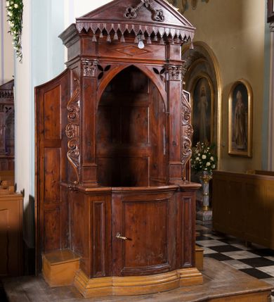 Zdjęcie nr 1: Drewniany konfesjonał ustawiony na jednostopniowym podwyższeniu na rzucie zbliżonym do prostokąta. Na profilowanym cokole drzwiczki wypukłe, wypełnione prostokątną płyciną, flankowane podobnymi, ale węższymi polami. Boki konfesjonału gładkie, przeprute otworami, z dostawionymi klęcznikami i zamknięte z obu stron wąskimi ściankami z płycinami, zakończonymi od góry skośnie. Baldachim z trójkątnym frontonem posadowiony na pilastrach korynckich dźwigających belkowanie dekorowane na środku leżącym rombem. Otwór zamknięty łukiem ostrym z trójliśćmi w przyłuczach, flankowany uszakami utworzonymi z liści i wolut odchodzących od centralnie ulokowanej rozety. Fronton wypukły, z umieszczonymi na środku skrzyżowanymi kluczami Piotrowymi, z rozetą na środku, od dołu dekorowany lambrekinem. Zaplecek dekorowany płycinami w kształcie leżących prostokątów. Struktura bejcowana na ciemnobrązowo. 