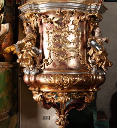 Zdjęcie nr 4: Ambona przyścienna, wisząca, z wejściem od tyłu dwubiegowymi schodami z podestem. Kosz na rzucie owalu o wklęsłych ściankach bocznych i wypukłej frontowej, na wypukłym cokole zdobionym od frontu płaskorzeźbionym motywem rocaille’u. U spodu kosza pięć kabłąków dekorowanych u góry palmetami i fragmentami rocaille’ów, zakończonych zwisem w formie winnego grona. Ścianki kosza opięte wklęsłym pilastrami; od frontu z figurami św. Jana i św. Mateusza siedzących na obłokach, trzymających otwarte księgi z napisem: OVI EX DEO EST VERBA DEI AUDIT / PROPTEREA VOS NON AUDITIS, QVIA EX DEO NON ESTIS. Joanne 8,47; oraz: ERAT ENIM / DOCENS EOS / SICUT PO=/TESTATEM /HABENS / ET NON / SICUT / SCRIBAE / EORUM / ET PHARI=/SAEI. Mat 7,18; na tle pozostałych pilastrów symbole ewangelistów: orzeł trzymający w ‘szponach kałamarz oraz głowa wołu. Ścianki w polach dekorowane rocaille’ami, frontowa z różanymi gałązkami. Kosz zwieńczony profilowanym gzymsem, występującym nad pilastrami. Na zaplecku obraz Szymona Kawalskiego Rozesłanie Apostołów, w kształcie leżącego prostokąta, w profilowanej, złoconej ramie, ujęty uszami utworzonymi z motywów rocaille’ów. Baldachim powtarza rzut kosza, wsparty na belkowaniu dekorowanym palmetami i rocaille’ami w partii fryzu, z podwieszonym lambrekinem. Powyżej ażurowa konstrukcja z wolut dekorowanych rocaille’ami, zbliżona do korony, otaczająca półpostać Chrystusa trzymającego jabłko królewskie i laskę pasterską. U spodu baldachimu podwieszona Gołębica Ducha Świętego w glorii. Struktura marmoryzowana laserunkowo, profile srebrzone, ornamenty złocone.