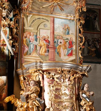 Zdjęcie nr 3: Ambona przyścienna, wisząca, z wejściem od tyłu dwubiegowymi schodami z podestem. Kosz na rzucie owalu o wklęsłych ściankach bocznych i wypukłej frontowej, na wypukłym cokole zdobionym od frontu płaskorzeźbionym motywem rocaille’u. U spodu kosza pięć kabłąków dekorowanych u góry palmetami i fragmentami rocaille’ów, zakończonych zwisem w formie winnego grona. Ścianki kosza opięte wklęsłym pilastrami; od frontu z figurami św. Jana i św. Mateusza siedzących na obłokach, trzymających otwarte księgi z napisem: OVI EX DEO EST VERBA DEI AUDIT / PROPTEREA VOS NON AUDITIS, QVIA EX DEO NON ESTIS. Joanne 8,47; oraz: ERAT ENIM / DOCENS EOS / SICUT PO=/TESTATEM /HABENS / ET NON / SICUT / SCRIBAE / EORUM / ET PHARI=/SAEI. Mat 7,18; na tle pozostałych pilastrów symbole ewangelistów: orzeł trzymający w ‘szponach kałamarz oraz głowa wołu. Ścianki w polach dekorowane rocaille’ami, frontowa z różanymi gałązkami. Kosz zwieńczony profilowanym gzymsem, występującym nad pilastrami. Na zaplecku obraz Szymona Kawalskiego Rozesłanie Apostołów, w kształcie leżącego prostokąta, w profilowanej, złoconej ramie, ujęty uszami utworzonymi z motywów rocaille’ów. Baldachim powtarza rzut kosza, wsparty na belkowaniu dekorowanym palmetami i rocaille’ami w partii fryzu, z podwieszonym lambrekinem. Powyżej ażurowa konstrukcja z wolut dekorowanych rocaille’ami, zbliżona do korony, otaczająca półpostać Chrystusa trzymającego jabłko królewskie i laskę pasterską. U spodu baldachimu podwieszona Gołębica Ducha Świętego w glorii. Struktura marmoryzowana laserunkowo, profile srebrzone, ornamenty złocone.